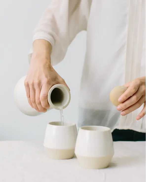 Person Pouring Beverage From Carafe Into Two Identical Cups While Holding Lid in Opposite Hand