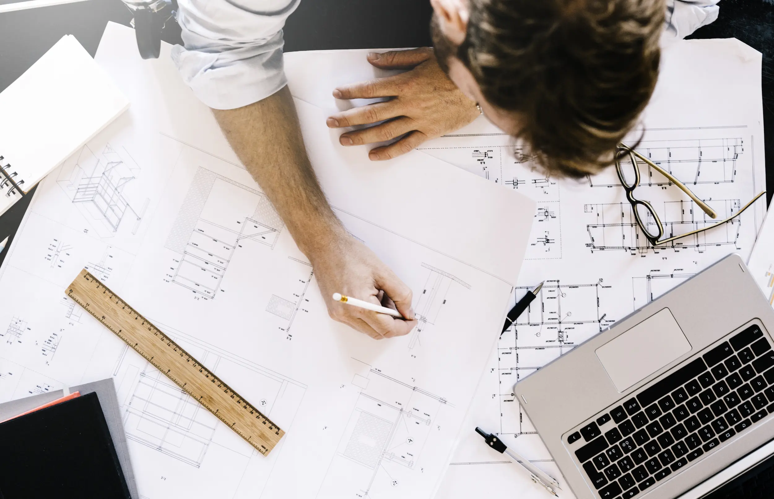 Birdseye View of Man Drawing Building Dimensions With Glasses, Ruler, and Laptop on the Table